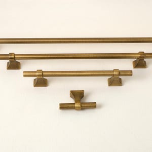 May include: A collection of four brass-toned metal cabinet pulls and a knob, each featuring a cylindrical bar design supported by two flared, square-based pedestals. The hardware displays a brushed metallic finish with a warm, golden-brown hue.