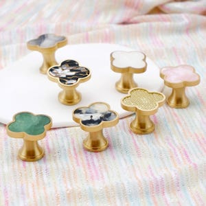 May include: A collection of decorative cabinet knobs with a cloverleaf design. The knobs feature various colors and patterns, including white, pink, green, and black, with gold-colored bases. These hardware accents add a touch of elegance to furniture.