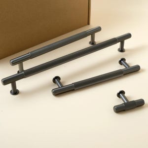 Puede incluir: Un juego de cuatro tiradores de armario de color gris oscuro con un diseño texturizado y acanalado. Dos tiradores más grandes y dos más pequeños están dispuestos sobre una superficie beige claro. Los tiradores tienen un diseño en forma de T y son de metal.