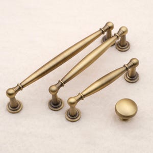 May include: Three antique brass cabinet pulls of varying lengths and one matching round cabinet knob. Each pull features a tapered center bar with rounded ends and two circular mounting bases. The finish is a brushed, muted metallic gold tone.