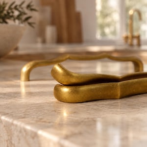 May include: Two curved, gold-toned metal cabinet pulls with a smooth, matte finish. The handles feature a gentle, undulating shape with rounded ends, positioned horizontally on a light-colored, textured stone surface in a bright, indoor setting.