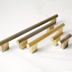Peut inclure: Une collection de poignées de meubles en laiton de différentes tailles. Les poignées ont un design cylindrique texturé et sont montées sur des bases rectangulaires. La finition en laiton varie du bronze foncé à l'or plus clair.