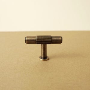 Pu&ograve; includere: Un pomello per mobili a forma di T con finitura in ottone anticato. La barra orizzontale presenta una sezione centrale con una fine texture a coste, affiancata da estremit&agrave; cilindriche lisce. Un perno verticale collega la barra a una base circolare.