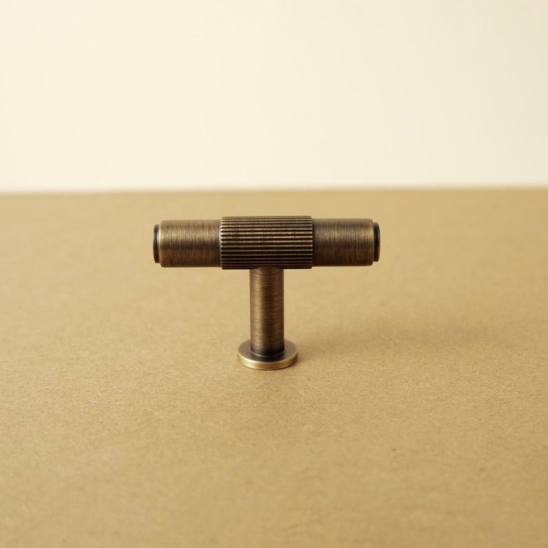 Pu&ograve; includere: Un pomello per mobili a forma di T con finitura in ottone anticato. La barra orizzontale presenta una sezione centrale con una fine texture a coste, affiancata da estremit&agrave; cilindriche lisce. Un perno verticale collega la barra a una base circolare.