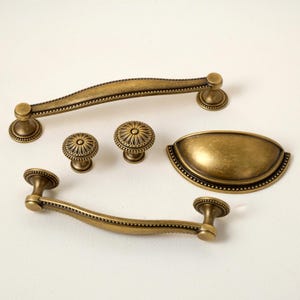 May include: An assortment of antique brass cabinet hardware, including two handles, two knobs, and a cup pull. The hardware features a detailed design with a warm, aged finish, perfect for kitchen or bathroom renovations.