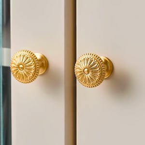 May include: Two gold-colored cabinet knobs with a floral design. The knobs are round with a detailed pattern and are mounted on a light-colored cabinet door. The knobs have a shiny finish and are centered in the image.