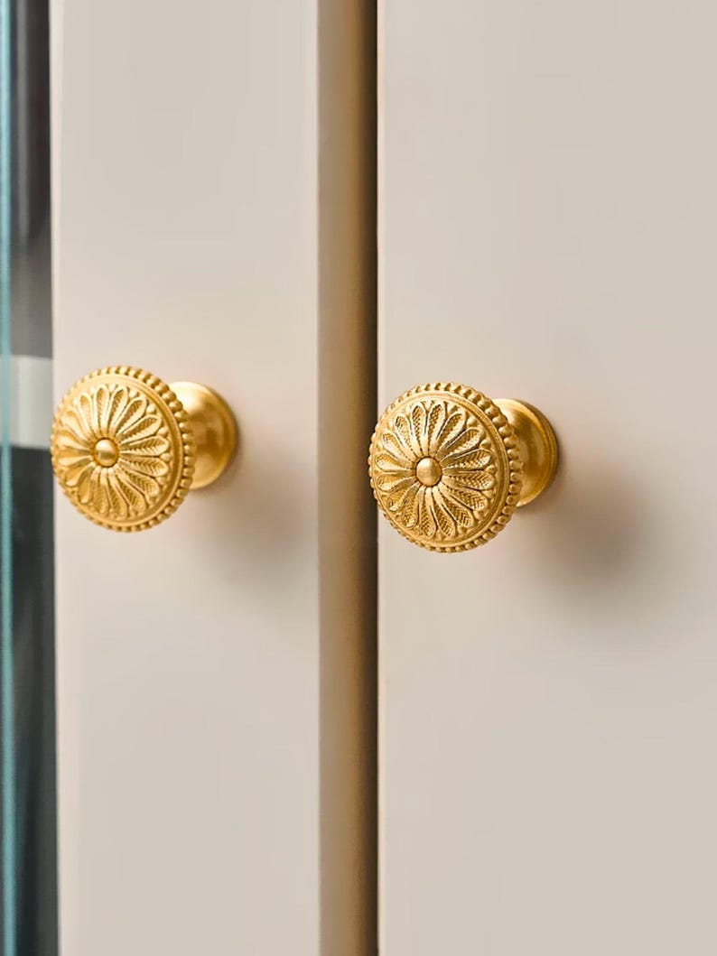 May include: Two gold-colored cabinet knobs with a floral design. The knobs are round with a detailed pattern and are mounted on a light-colored cabinet door. The knobs have a shiny finish and are centered in the image.