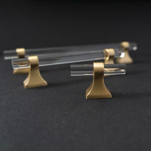 May include: Clear acrylic cabinet pulls with brass-coloured bases. The pulls have a cylindrical design and are set against a dark background. The hardware is designed for furniture and home decor.