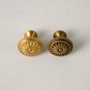 May include: Two antique brass cabinet knobs. One knob features a sunburst design, while the other has a floral pattern with a beaded border. Both knobs have a rounded base and are made of a gold-toned metal.