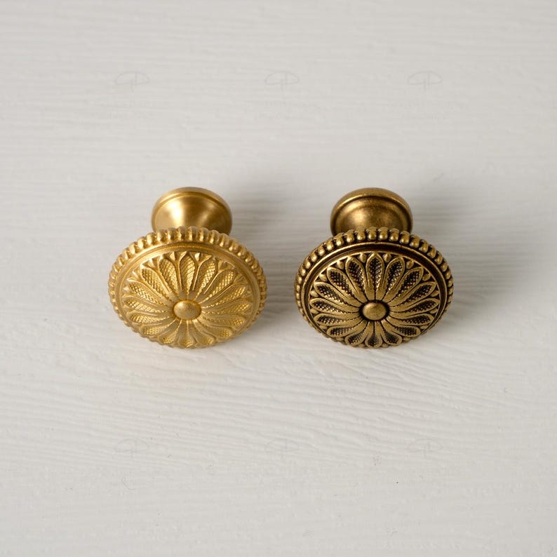 May include: Two antique brass cabinet knobs. One knob features a sunburst design, while the other has a floral pattern with a beaded border. Both knobs have a rounded base and are made of a gold-toned metal.