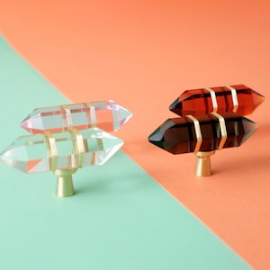 Puede incluir: Cuatro pomos de cristal para armarios con detalles dorados. Dos son transparentes con bandas doradas, y dos son ámbar y negros con bandas doradas. Los pomos tienen forma facetada de gema y están montados sobre bases doradas. El fondo está dividido diagonalmente con naranja y verde.