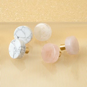 May include: A selection of cabinet knobs in different colours and patterns. The knobs include white marble with grey veining, translucent white, and pink quartz. Each knob has a gold-coloured base.