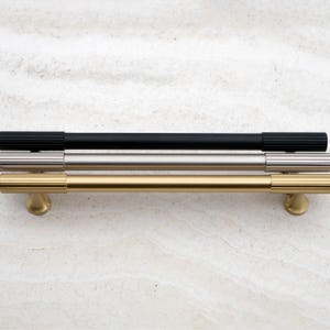 K&ouml;nnte beinhalten: Drei Schrankgriffe in verschiedenen Ausf&uuml;hrungen: Schwarz, Silber und Gold. Der schwarze Griff hat eine gerippte Textur an den Enden. Der silberne Griff ist glatt. Der goldene Griff hat eine gerippte Textur und einen runden Sockel.