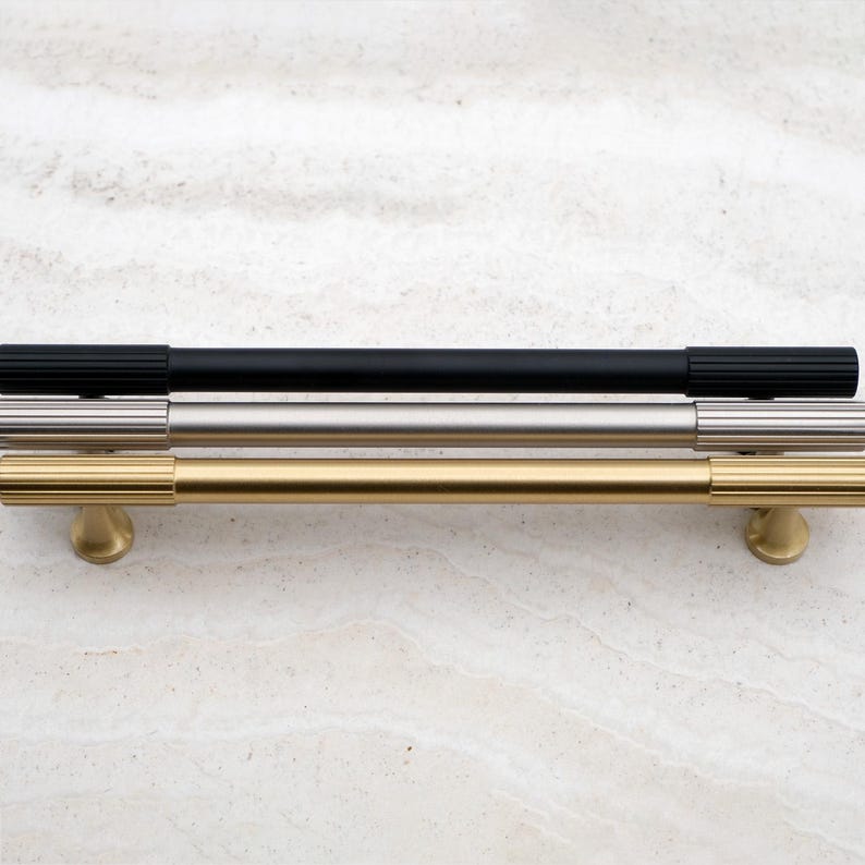 K&ouml;nnte beinhalten: Drei Schrankgriffe in verschiedenen Ausf&uuml;hrungen: Schwarz, Silber und Gold. Der schwarze Griff hat eine gerippte Textur an den Enden. Der silberne Griff ist glatt. Der goldene Griff hat eine gerippte Textur und einen runden Sockel.