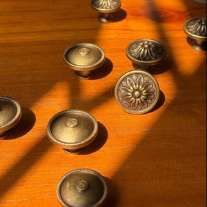 Puede incluir: Una colección de pomos redondos para armarios hechos de metal con tono de latón antiguo. Algunos pomos presentan un patrón floral detallado en relieve en la cara, mientras que otros tienen una superficie lisa con un pequeño detalle circular central.