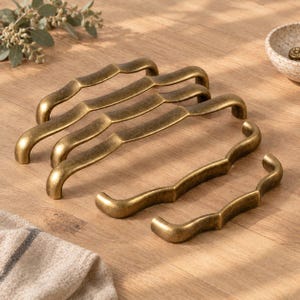 May include: A set of four antique brass cabinet pulls arranged in a cascading pattern. Each handle features a distinct wavy, curved silhouette with a weathered, metallic finish. The pulls vary in length, showcasing a consistent design aesthetic.