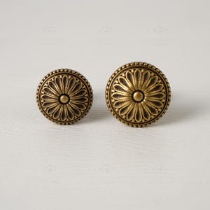 May include: Two antique brass cabinet knobs with a floral design. The knobs are round with a detailed pattern of petals radiating from a central point. The edges are adorned with small, raised beads. The knobs are displayed on a white surface.