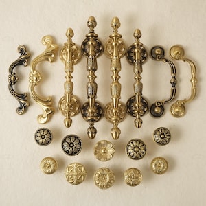 Puede incluir: Una colección de herrajes de gabinete de latón antiguo, que incluye manijas ornamentadas y perillas decorativas. Las manijas presentan intrincados diseños florales y de volutas, con acabados dorados y negros. Las perillas tienen varios patrones florales.