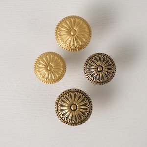 May include: Four decorative cabinet knobs in gold and bronze tones. Each knob features a central floral design surrounded by a beaded edge. The knobs are arranged on a white surface, showcasing their intricate details.