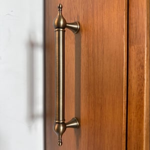 May include: A vertical cabinet handle with an antique brass finish, featuring a cylindrical center bar and decorative finials at both ends. The hardware is mounted onto a smooth, medium-toned wooden surface with a visible vertical grain pattern.