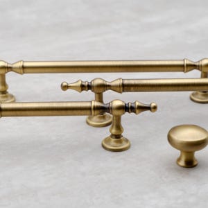 May include: A collection of antique brass cabinet hardware including two long horizontal drawer pulls with decorative finials and one round mushroom-shaped cabinet knob. Each piece features a brushed metallic finish with dark accents.