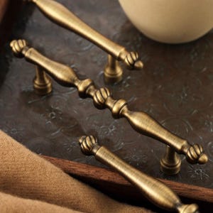 May include: Three antique brass cabinet pulls featuring a central spindle design with rounded, ribbed accents at the ends and middle. Each handle is mounted on two small, circular pedestals, showcasing a brushed metallic finish with dark highlights.