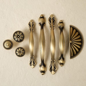 May include: A collection of antique brass cabinet hardware, including knobs, pulls, and a half-round backplate. The knobs have a floral design, while the pulls are elongated with decorative detailing. The hardware has a vintage aesthetic.