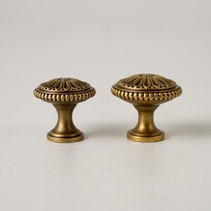May include: Two antique brass cabinet knobs. Each knob features a detailed, floral-inspired design on the round top and a sturdy, pedestal-style base. The knobs are a warm, golden color, suggesting an aged finish, and are set against a white background.