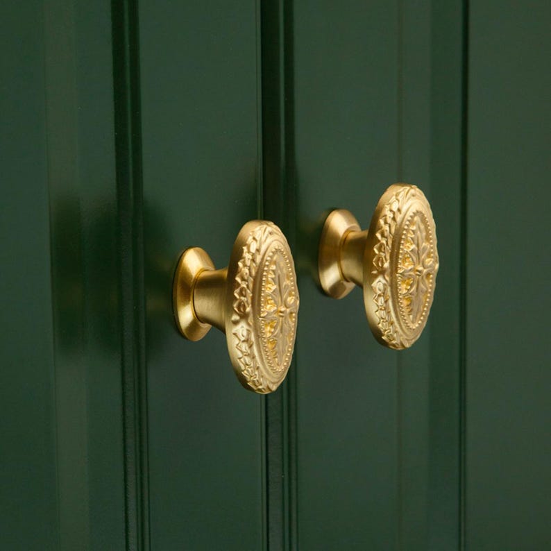 Puede incluir: Primer plano de dos pomos de armario ovalados dorados con detalles florales ornamentados. Los pomos est&aacute;n montados en una puerta de armario verde oscuro. Los pomos tienen una est&eacute;tica vintage.