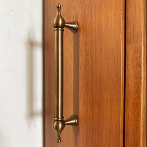 May include: A vertical antique brass cabinet handle with a cylindrical center and decorative finials at both ends. The handle features a brushed metallic finish and is mounted onto a smooth, light brown wooden surface with a visible grain pattern.