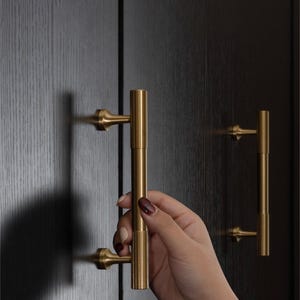 K&ouml;nnte beinhalten: Nahaufnahme von goldfarbenen Schrankgriffen mit vertikalem, zylindrischem Design. Die Griffe sind an einer dunklen Holzschrankt&uuml;r angebracht. Das Bild zeigt eine Hand, die einen der Griffe h&auml;lt. Die Hardware hat eine klassische, elegante &Auml;sthetik.