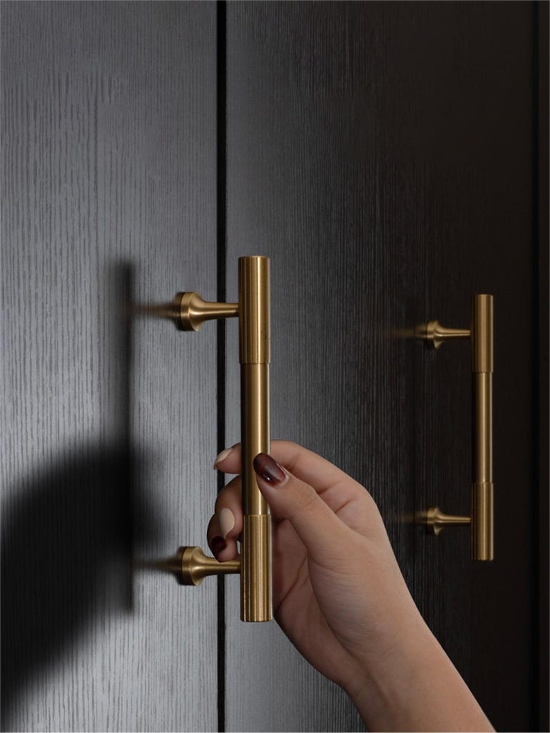 K&ouml;nnte beinhalten: Nahaufnahme von goldfarbenen Schrankgriffen mit vertikalem, zylindrischem Design. Die Griffe sind an einer dunklen Holzschrankt&uuml;r angebracht. Das Bild zeigt eine Hand, die einen der Griffe h&auml;lt. Die Hardware hat eine klassische, elegante &Auml;sthetik.
