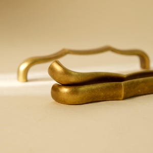 May include: Close-up of two gold-colored metal cabinet pulls. One is a pair of tongs, and the other is a curved handle. The cabinet hardware has a matte finish and a simple, elegant design. The background is a neutral beige.