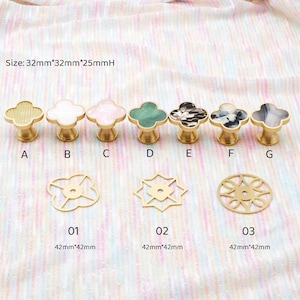 Op de afbeelding: Een verzameling decoratieve kastknoppen en -grepen in verschillende ontwerpen en kleuren. De knoppen hebben klavervormige toppen met goudkleurige bases. De afbeelding bevat ook goudkleurige decoratieve accenten. De tekst geeft de grootte van de knoppen aan.