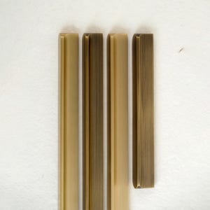 May include: Four vertical metal cabinet pulls in varying shades of gold and bronze. The pulls have a rectangular shape with rounded edges and are arranged against a white background. The finish appears to be brushed or aged.