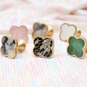 Op de afbeelding: Een verzameling decoratieve kastknoppen in een klaverbladontwerp. De knoppen hebben verschillende kleuren en patronen, waaronder roze, wit, groen en zwart-wit, met goudkleurige bases. Ideaal om een vleugje elegantie aan meubels toe te voegen.