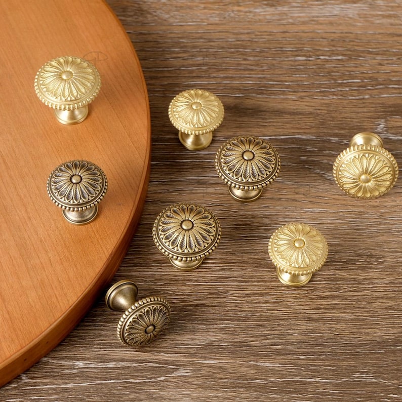 May include: A collection of antique-style cabinet knobs in various designs. The knobs are made of brass and feature intricate floral patterns. Some knobs are round, while others have an oval shape. The knobs are displayed on a wooden surface.