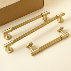 May include: Three gold-toned metal cabinet pulls with a fluted, cylindrical design. Each handle features a ribbed center section and two circular mounting bases at the ends, providing a consistent metallic finish across all three hardware pieces.