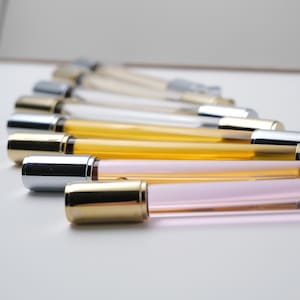 May include: An assortment of roll-on perfume bottles with clear, amber, and pink glass tubes. The bottles have metallic gold and silver caps. The bottles are arranged diagonally on a white surface.