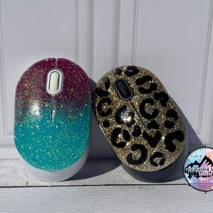 May include: Two wireless computer mice. One mouse has a glittery gradient design, transitioning from purple to teal. The other mouse features a gold glitter leopard print pattern with black spots. Both have a scroll wheel and buttons.