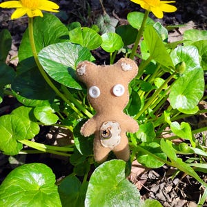 Puede incluir: Un oso de peluche marrón hecho a mano con ojos de botón y un parche decorativo en el vientre, en medio de un follaje verde vibrante y flores amarillas brillantes. Un artículo encantador y artesanal.