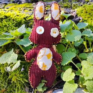 Könnte beinhalten: Ein handgefertigtes, weinrotes Plüschkaninchen mit Knopfaugen und einem blütenförmigen Knopf. Die Ohren und der Bauch des Kaninchens weisen einen weißen Stoff mit gelbem Blumenmuster auf. Das Kaninchen ist von grünem Laub umgeben.