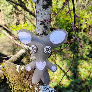 Puede incluir: Un ratón de fieltro gris hecho a mano con ojos de botón, orejas blancas y un pecho en forma de corazón rosa. El ratón cuelga de una rama de árbol, con un fondo natural verde y marrón.
