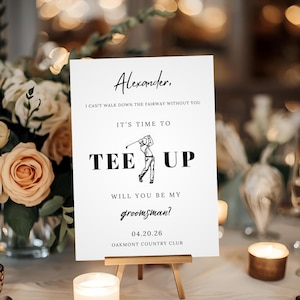 以下が含まれることがあります： 小さな木製イーゼルに立てられた白い看板。テキスト「アレクサンダー、あなたなしではフェアウェイを歩けません。ティーアップの時間です。私の新郎付添人になってくれますか？」が書かれ、花の装飾とキャンドルに囲まれています。