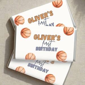 Op de afbeelding: Witte snoepreepverpakkingen met basketbalillustraties en de tekst "Oliver's First Birthday" in bruin en blauw. De verpakkingen zijn versierd met oranje basketballen.