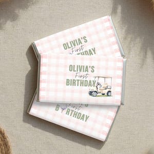 Pode incluir: Duas embalagens retangulares de barras de chocolate com um padrão xadrez rosa claro e branco. O texto diz OLIVIA'S First BIRTHDAY em letras verdes. Uma pequena ilustração de um carrinho de golfe e uma bola de golfe estão impressas.