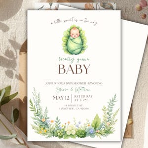 Może przedstawiać: Zaproszenie na baby shower z akwarelowymi ilustracjami. Zaproszenie przedstawia dziecko owinięte w zielony kocyk, otoczone kwiatowym wieńcem. Tekst zawiera "a little sprout is on the way" i "locally grown BABY".