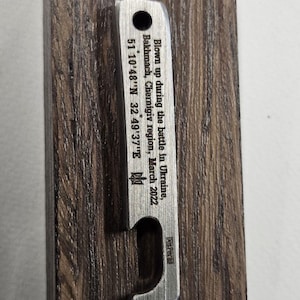 Puede incluir: Un abridor de botellas plateado con un extremo curvado y un agujero en la parte superior, montado en una placa de madera oscura. El abridor está grabado con texto sobre una batalla en Ucrania, incluyendo coordenadas y la fecha de marzo de 2022.