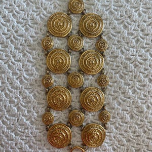 Peut inclure: Un bracelet en métal doré composé d'une série de médaillons circulaires interconnectés avec un motif de corde en spirale texturé. La pièce comprend un fermoir à bascule en haut et un anneau circulaire en bas, reliés par des anneaux métalliques.