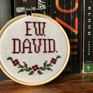 May include: A cross-stitch embroidery hoop with the phrase "EW, DAVID" in burgundy letters. A floral garland with burgundy flowers and green leaves decorates the bottom. The hoop is set against a backdrop of books.